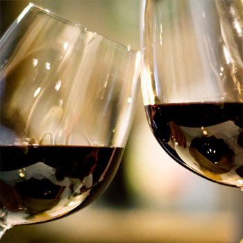 A closeup of 2 Bordeaux shaped glasses of red wine clinking cheers together.