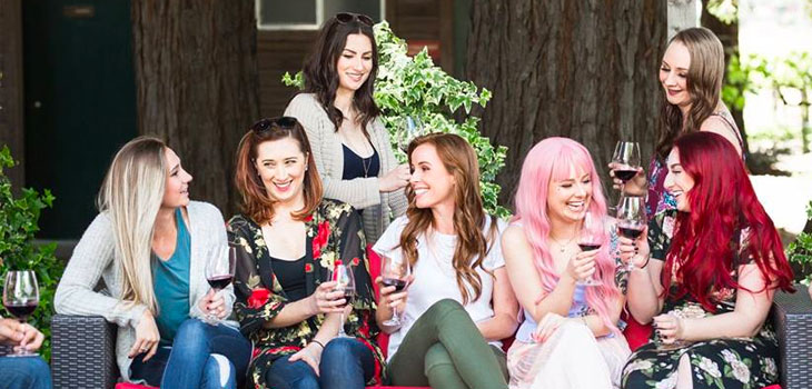 A group of 7 lively women in bright outfits sit on a patio couch smiling and laughing during a wine tasting in Sonoma at Trentadue.