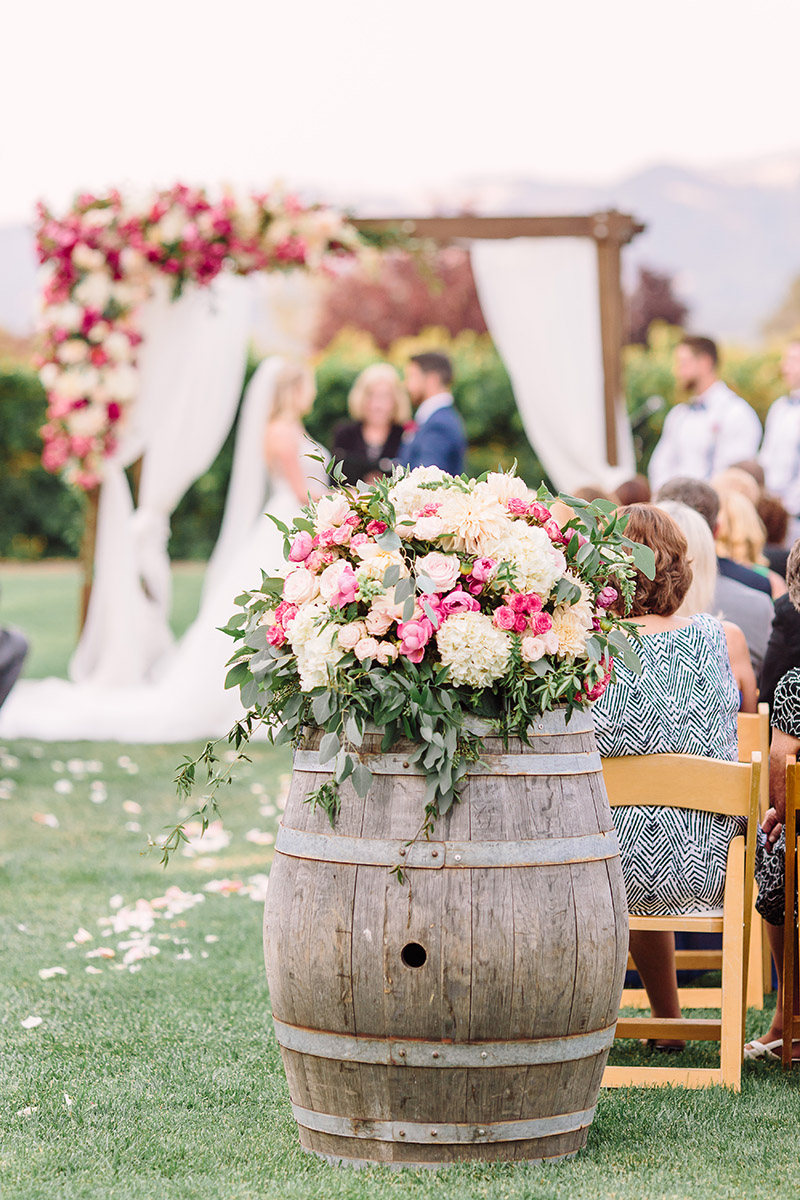 A closeup of an oak barrel decorated with a bouquet of pink flowers and greenery. A wine country wedding ceremony is taking place in the background.