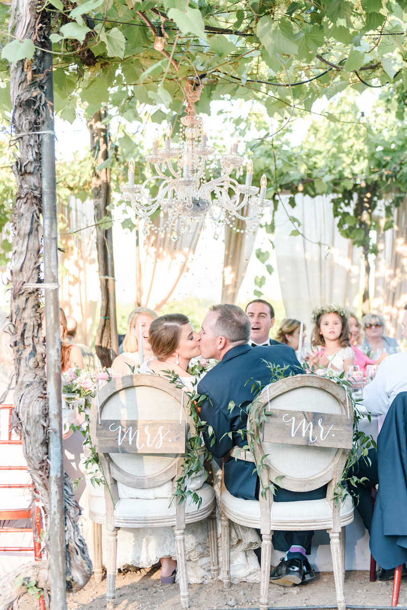 A rear view angle of a bride and groom kissing at a banquet table as their guests smile and look on. They sit beneath a decorated grape arbor at a Sonoma wedding venue with chairs that read Mr. and Mrs.