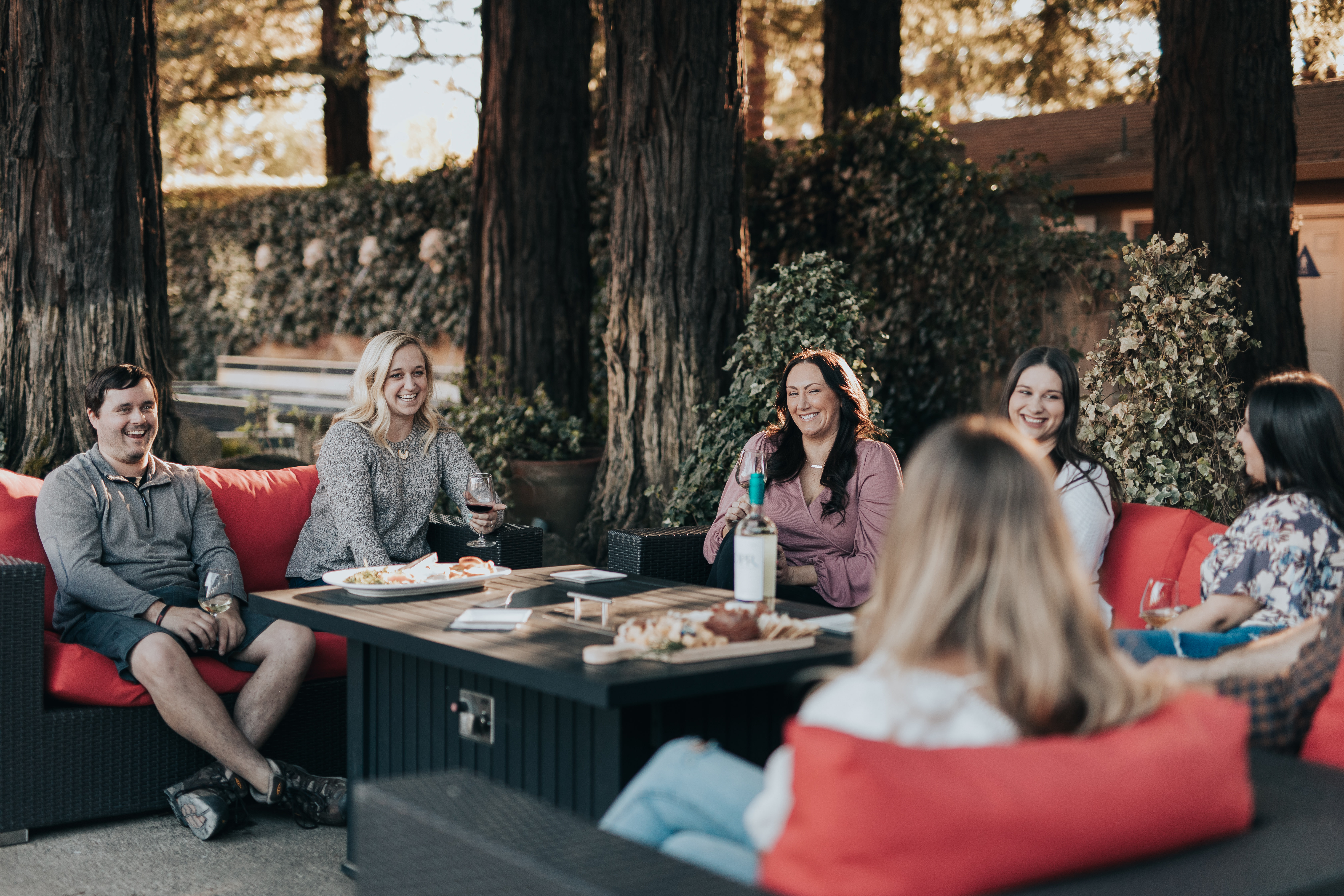 A group of people enjoying wine on red couches outdoors with trees and greenery in the background.