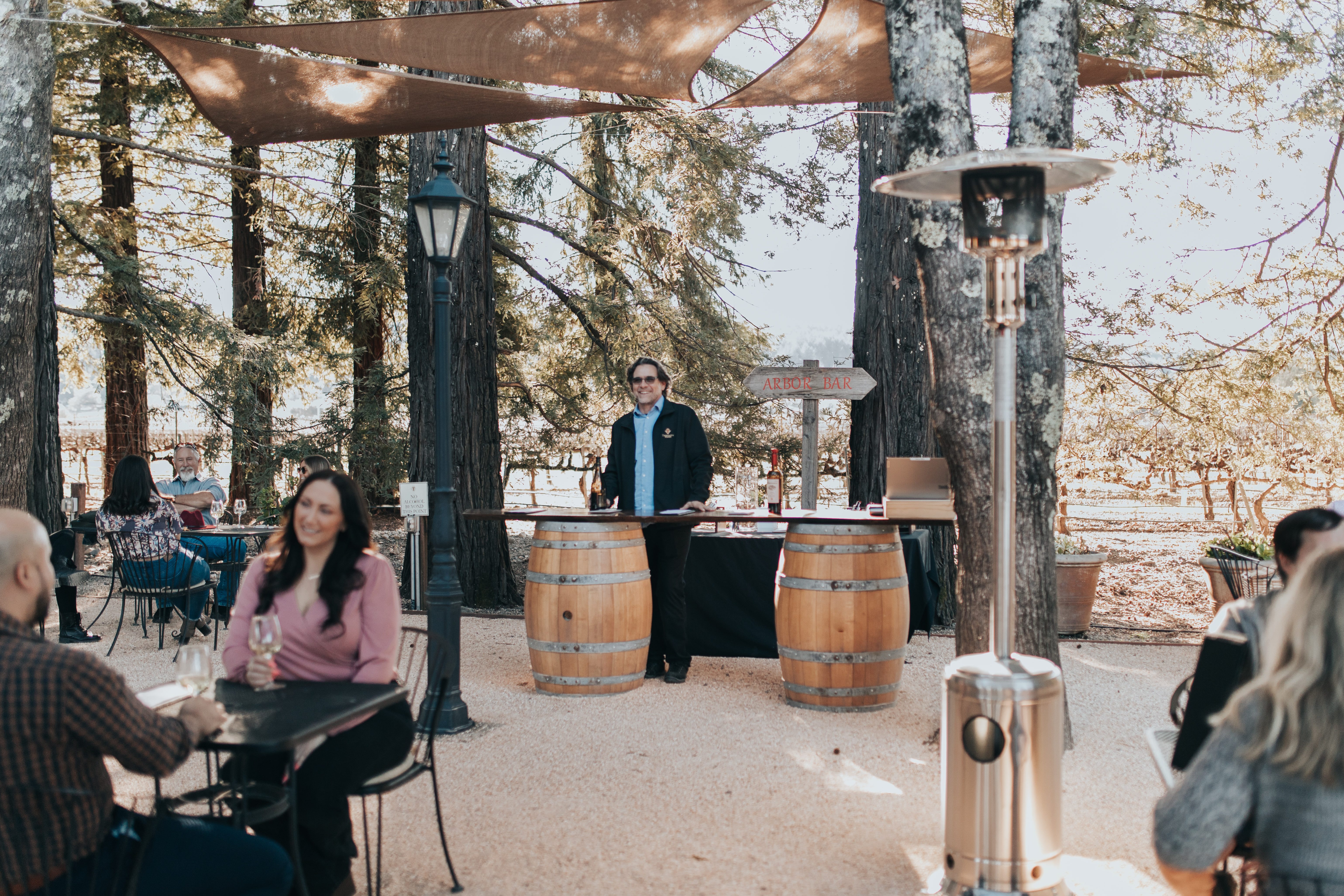 A man standing behind a bar made out of wine barrels holding a bottle of wine surrounded people seated at tables drinking wine.
