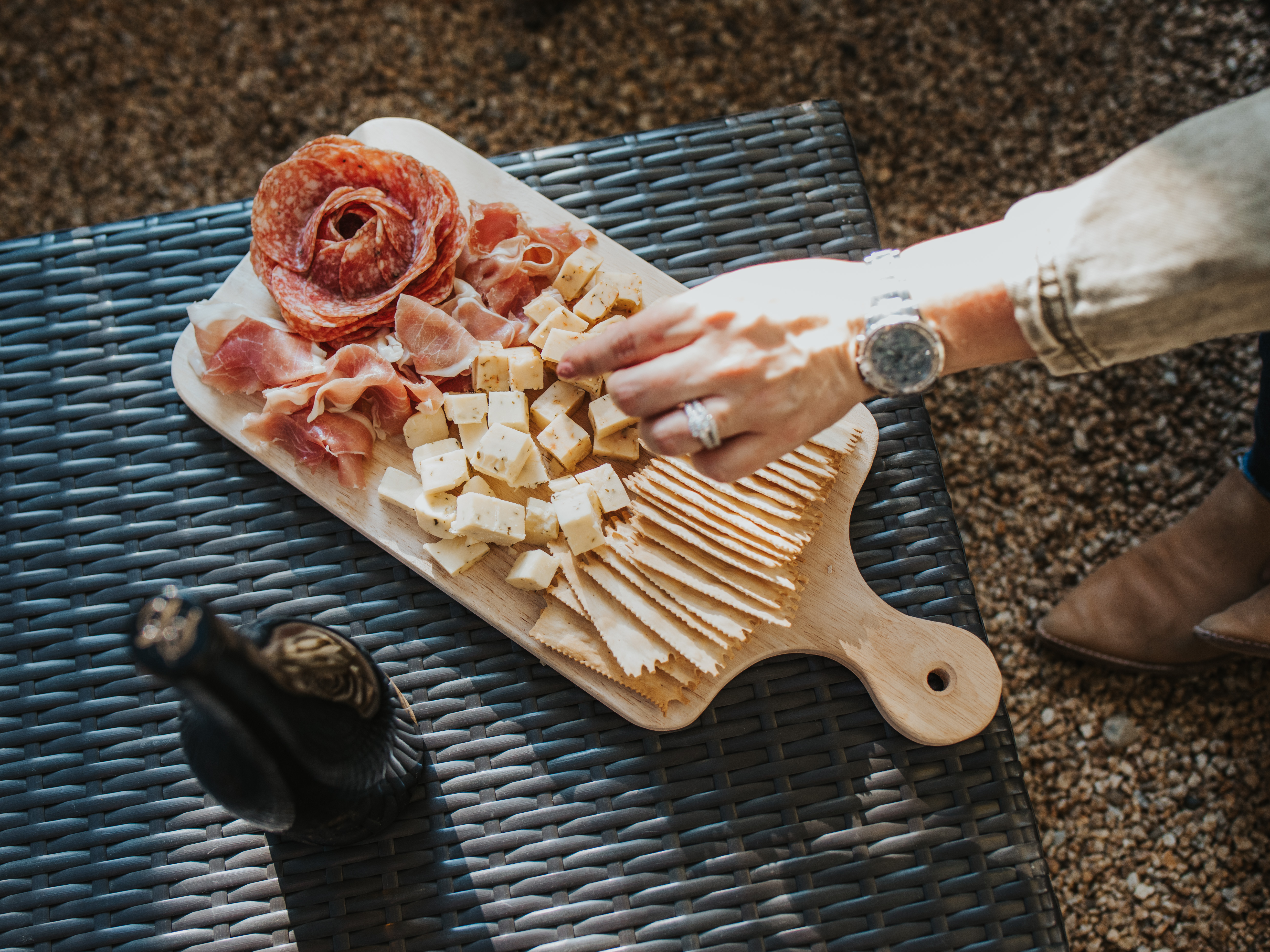 cubed cheese, salami and crackers on a wooden board with a bottle of wine out of focus and a hand reaching to grab cheese