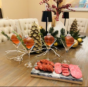 cocktail in christmas ornament in a martini glass served with sliced meats and holiday decorations in the background